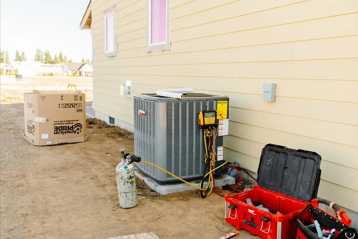 Outdoor HVAC unit with service gauges and tools beside yellow house siding